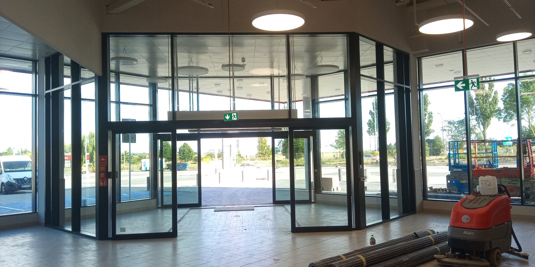 Szeroki kadr na szklane drzwi wejściowe do budynku z widocznym znakiem ewakuacyjnym, sprzętem budowlanym i rurami na podłodze z płytek.