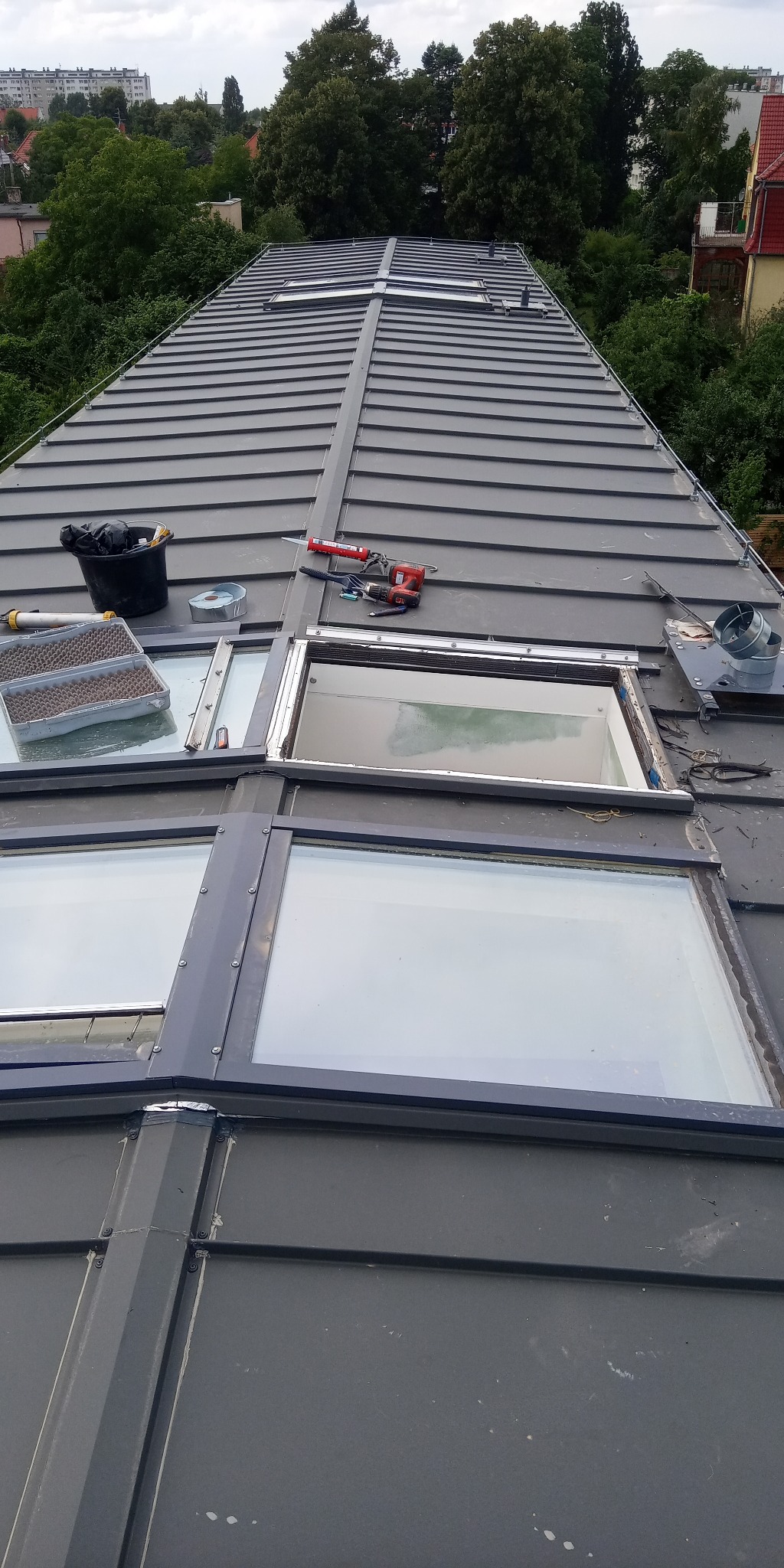 Montaż okien dachowych na metalowym dachu, widoczne narzędzia, pistolet do silikonu, puszka z pianką montażową i części okna.