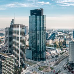 Kancelaria Finansowo - Ubezpieczeniowa Anna Gomulak - Panorama Warszawy z wieżowcami, w tym budynek Warty. Widok z góry na skrzyżowanie, zieleń i inne budynki w tle, przy słonecznej pogodzie.