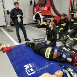 Instruktor demonstruje użycie worka ambu podczas symulacji akcji ratunkowej z udziałem strażaków i manekinów na tle wozu strażackiego.