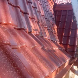Błyszczący dach z czerwonej dachówki ceramicznej, widok z dołu na krawędź dachu z zamontowaną rynną, fragment drabiny po prawej.