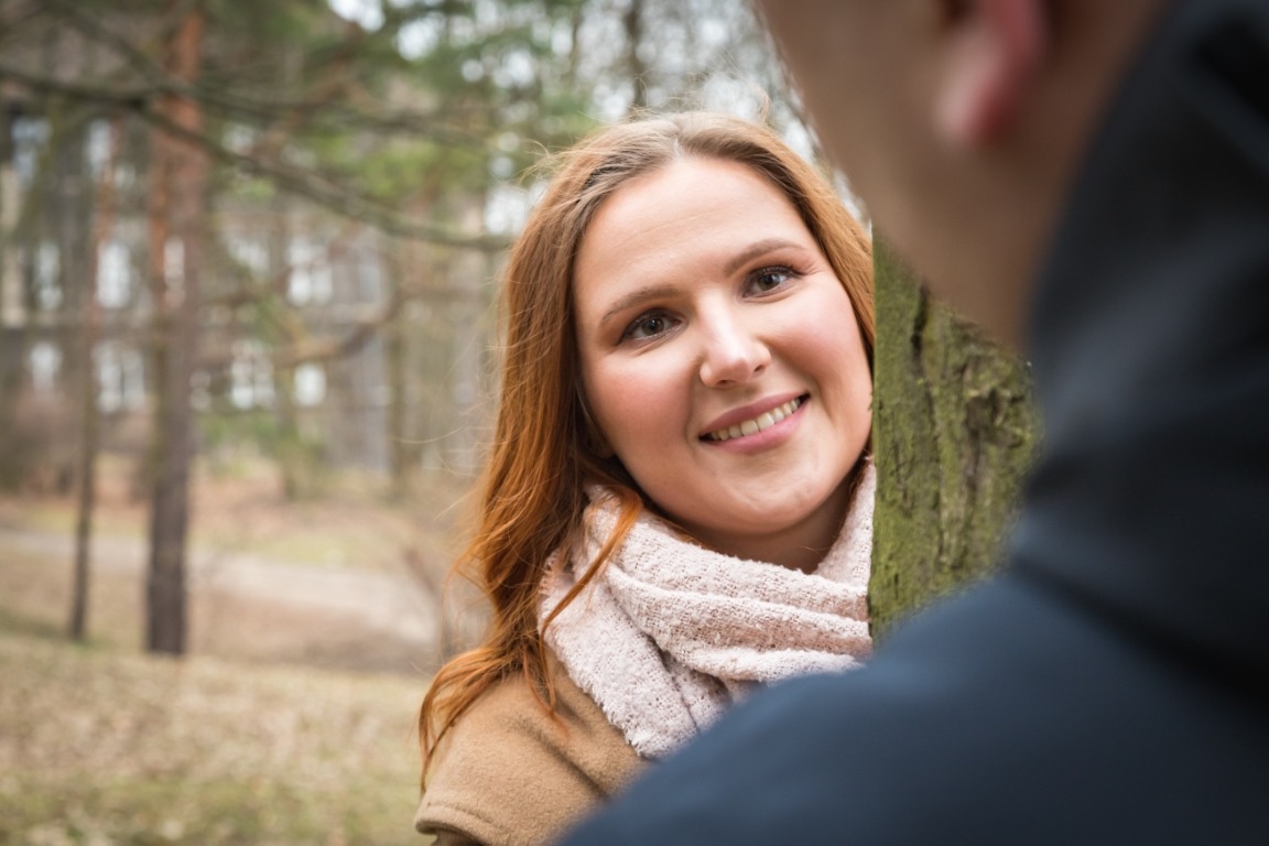 Uśmiechnięta kobieta z rudymi włosami i beżowym szalikiem, ukryta za drzewem w parku, z widocznym fragmentem sylwetki mężczyzny w ciemnej kurtce na pierwszym planie.