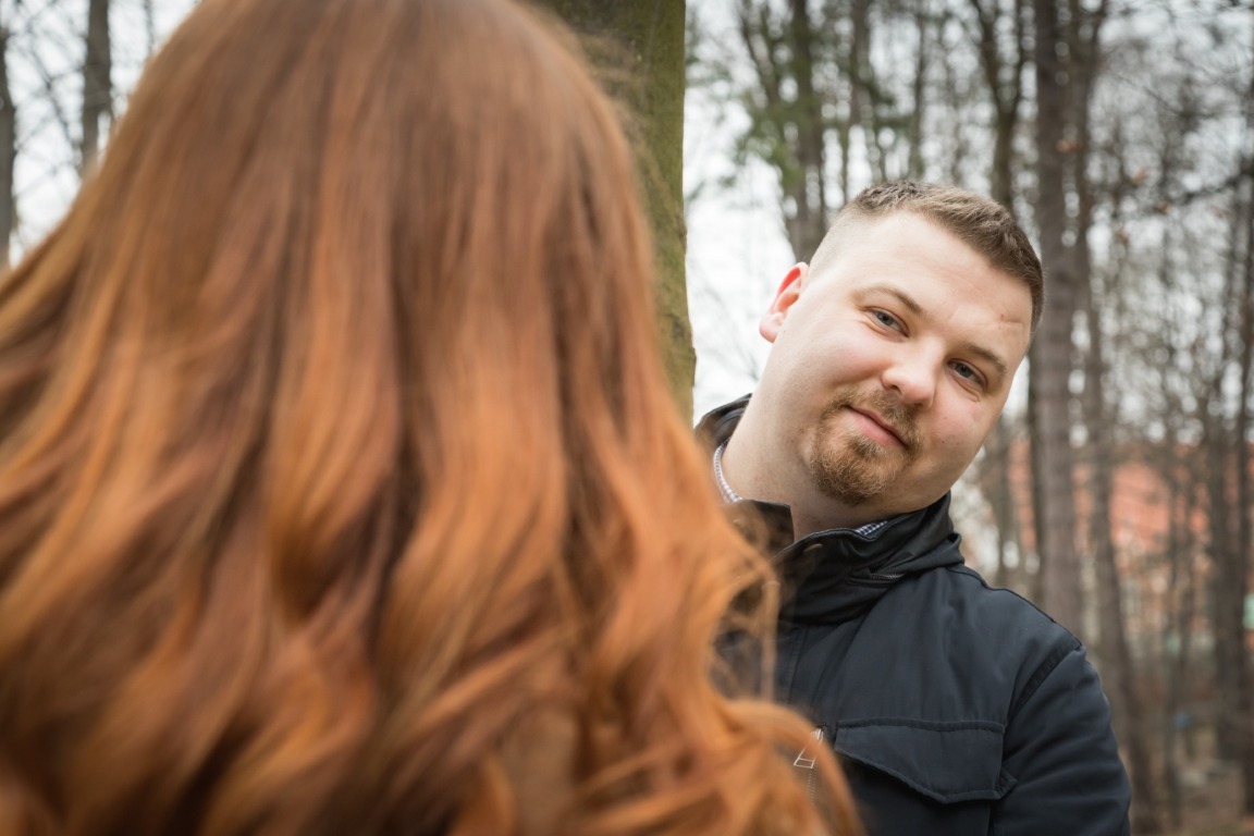 Mężczyzna w czarnej kurtce spogląda zza drzewa na kobietę z rudymi włosami podczas sesji zdjęciowej w plenerze.