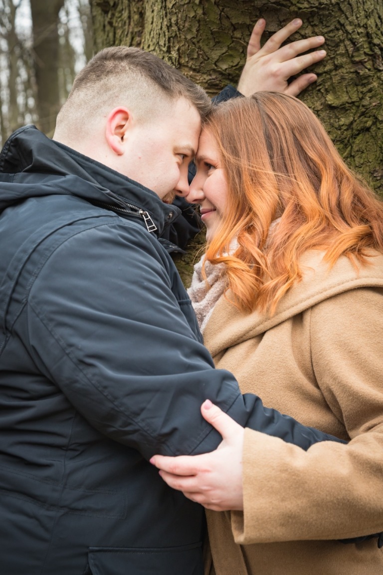 Zakochana para opiera czoła o siebie, stojąc przy drzewie w parku, kobieta ma rude włosy i beżowy płaszcz, mężczyzna w ciemnej kurtce.