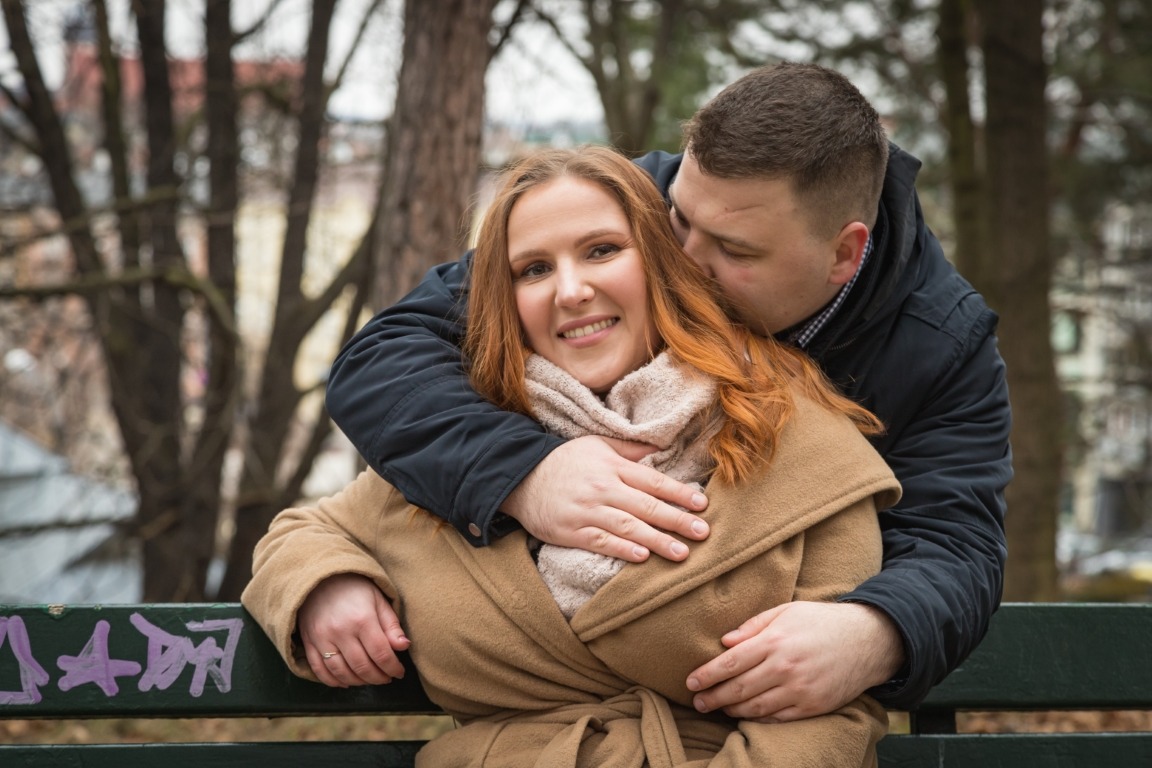 Uśmiechnięta kobieta w beżowym płaszczu, obejmowana przez mężczyznę na ławce w parku, z widocznym graffiti na oparciu ławki.
