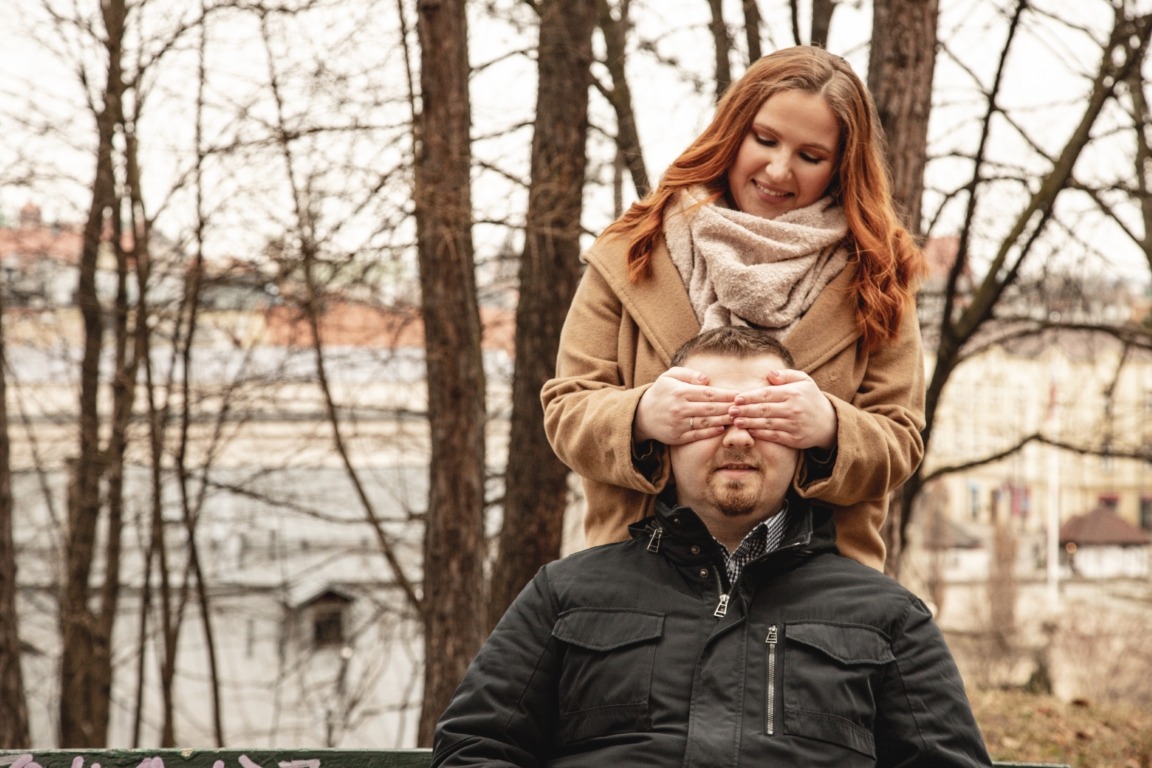 Uśmiechnięta ruda kobieta w beżowym płaszczu zakrywa oczy siedzącemu mężczyźnie w czarnej kurtce na tle parku z drzewami i budynkami.