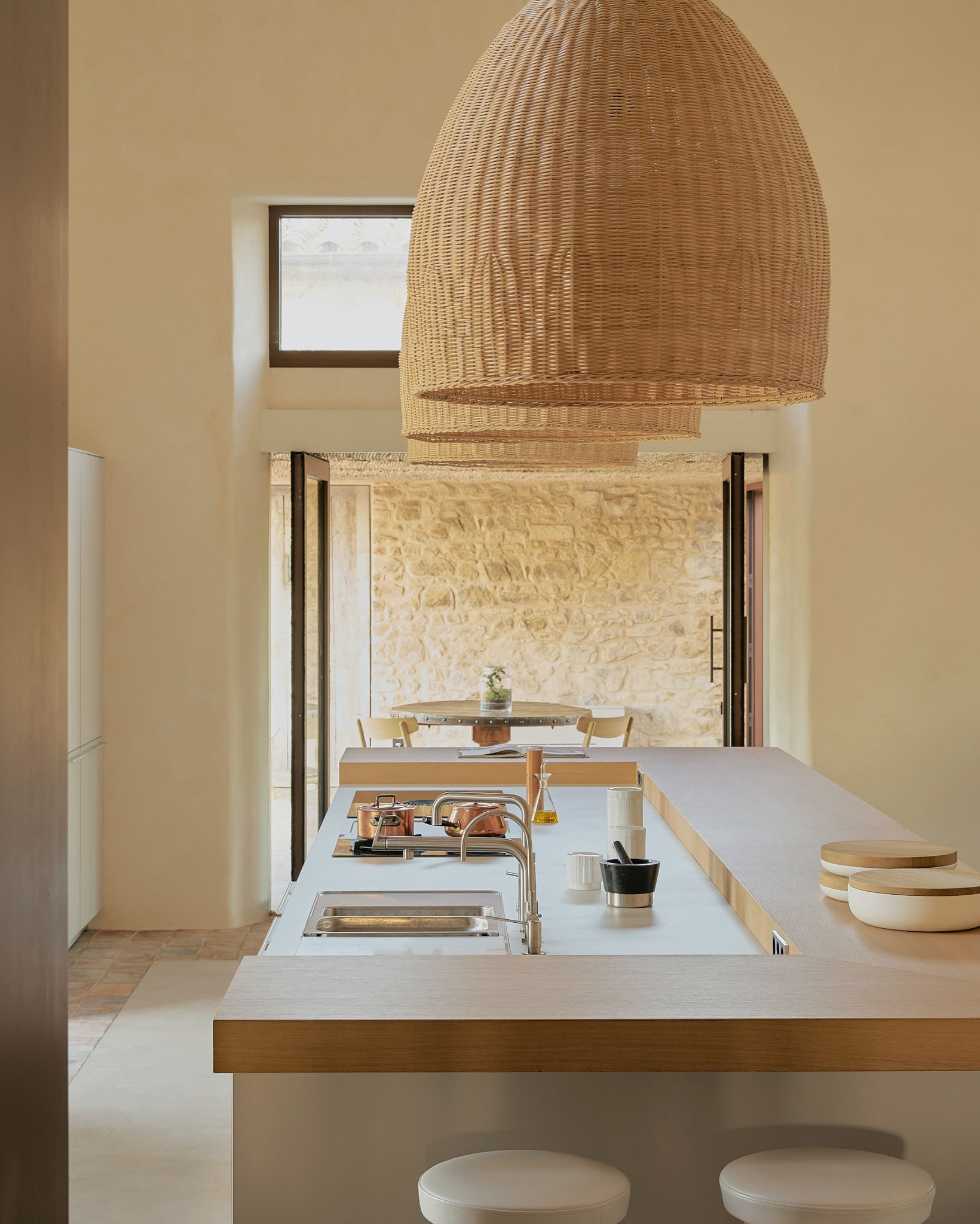 Jasna kuchnia z wyspą i wiklinowymi lampami, widok na kamienną ścianę i stół z krzesłami w tle, minimalistyczny styl.