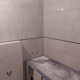 Montaż pionowych, białych płytek w kształcie migdałów na ścianie łazienki, widoczna zabudowa wanny w trakcie prac wykończeniowych, zasilikonowane krawędzie wanny.