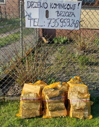 Worki z pociętym drewnem kominkowym brzozowym, ustawione na trawie przed siatką ogrodzeniową, na której wisi kartka z napisem 'Drzewo kominkowe! Brzoza TEL: 735 958 248'.