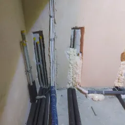 Wewnętrzne rury instalacyjne w trakcie montażu, częściowo zabezpieczone pianką montażową w ścianie, widoczne czarne izolacje termiczne i niebieski karbowany przewód ochronny.