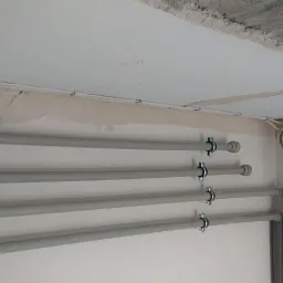 Szare rury instalacyjne zamocowane do sufitu za pomocą metalowych uchwytów, widoczne przewody elektryczne oraz fragment surowego sufitu i ściany w trakcie remontu.