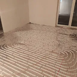 Białe rurki ogrzewania podłogowego ułożone na folii z czerwoną siatką, widoczne w surowym stanie pomieszczenia z oknem balkonowym.