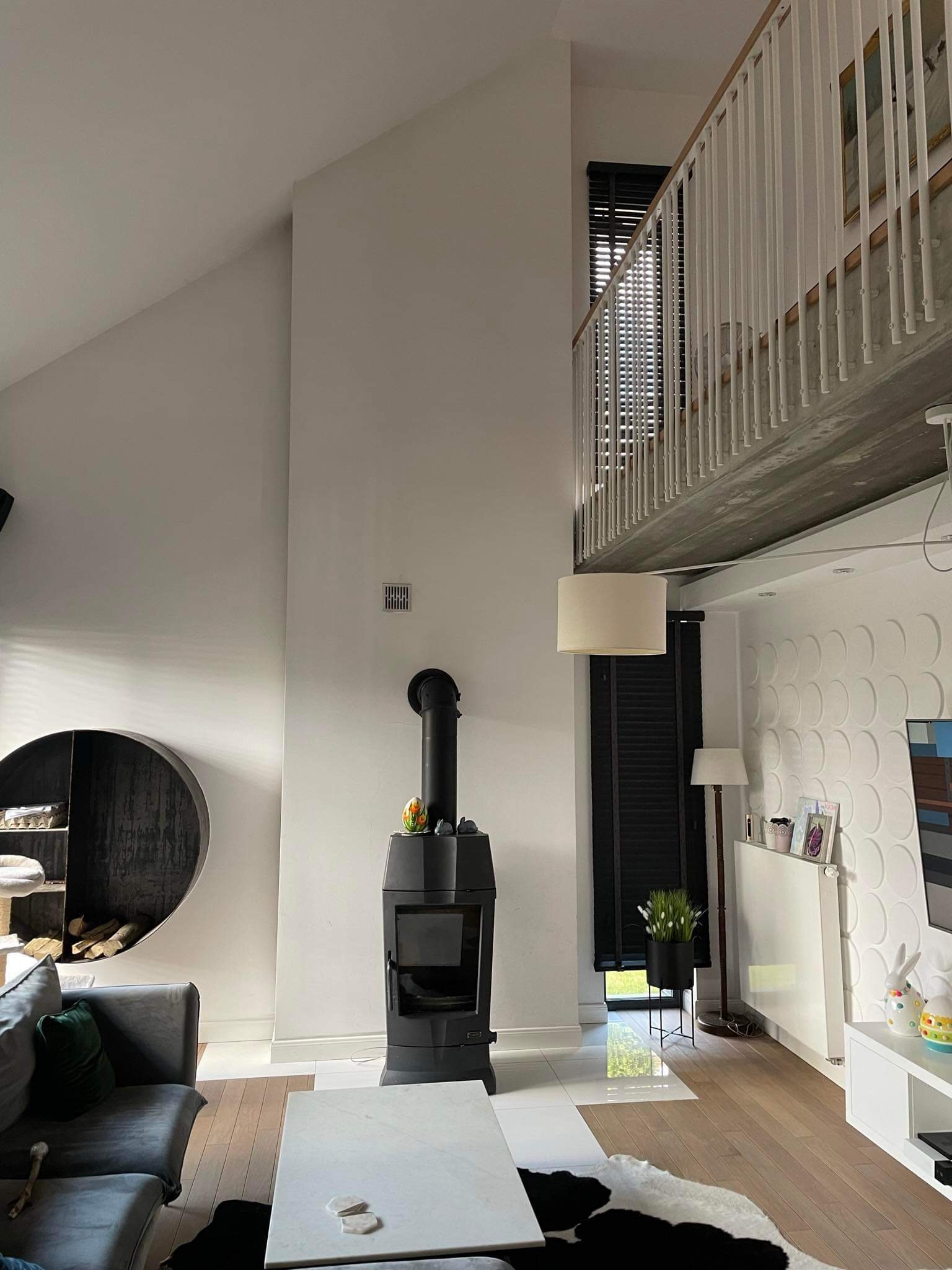 Wysoki salon z antresolą, czarnym piecem wolnostojącym, okrągłą półką na drewno i sofą w odcieniach szarości, oświetlony naturalnym światłem.