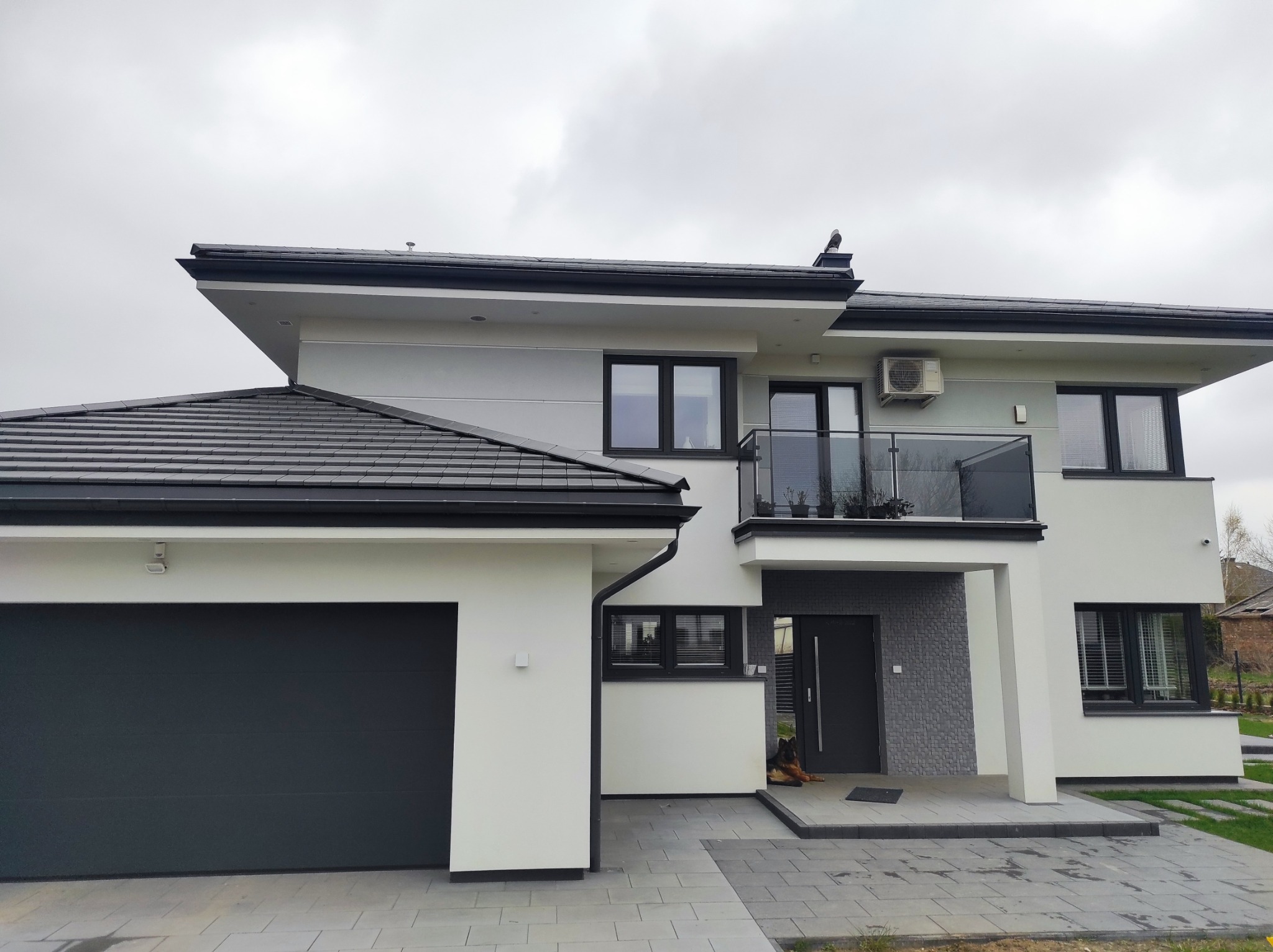 Nowoczesny dom z dwuspadowym dachem pokrytym ciemną dachówką, białymi ścianami, czarnymi oknami i garażem po lewej stronie; widoczny fragment balkonu z szklaną balustradą i klimatyzator na elewacji...