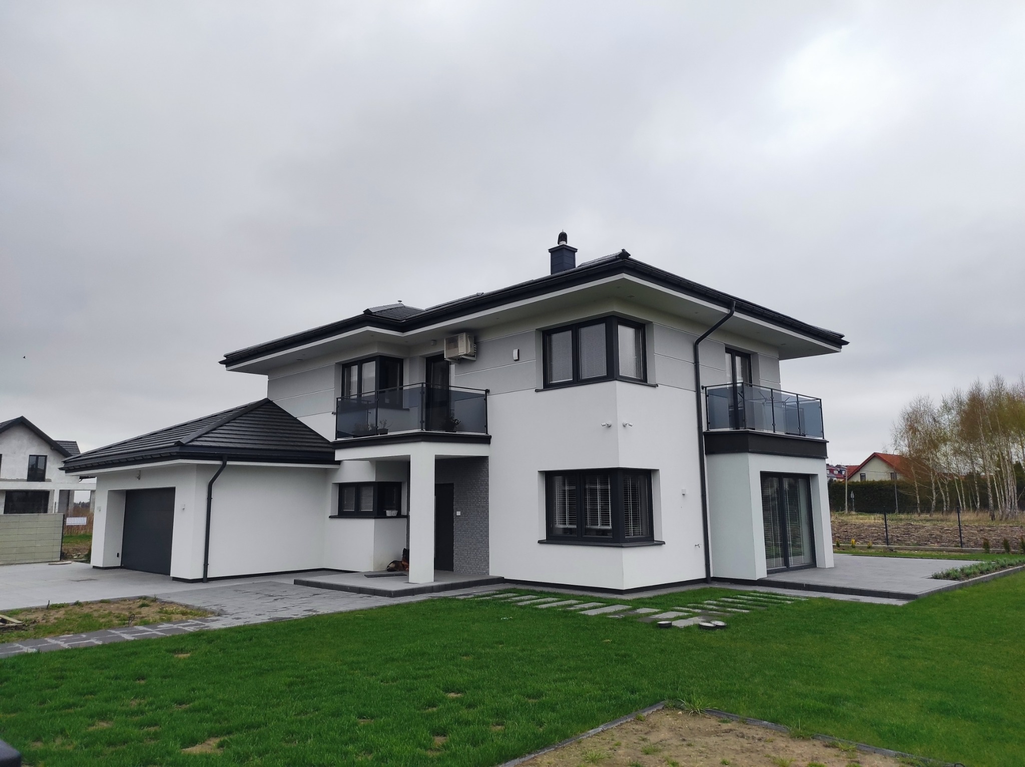 Nowoczesny dom jednorodzinny z płaskim dachem i balkonami z szklaną balustradą, biało-szara elewacja, czarna stolarka okienna i dachówka, trawnik z kamiennymi płytami.