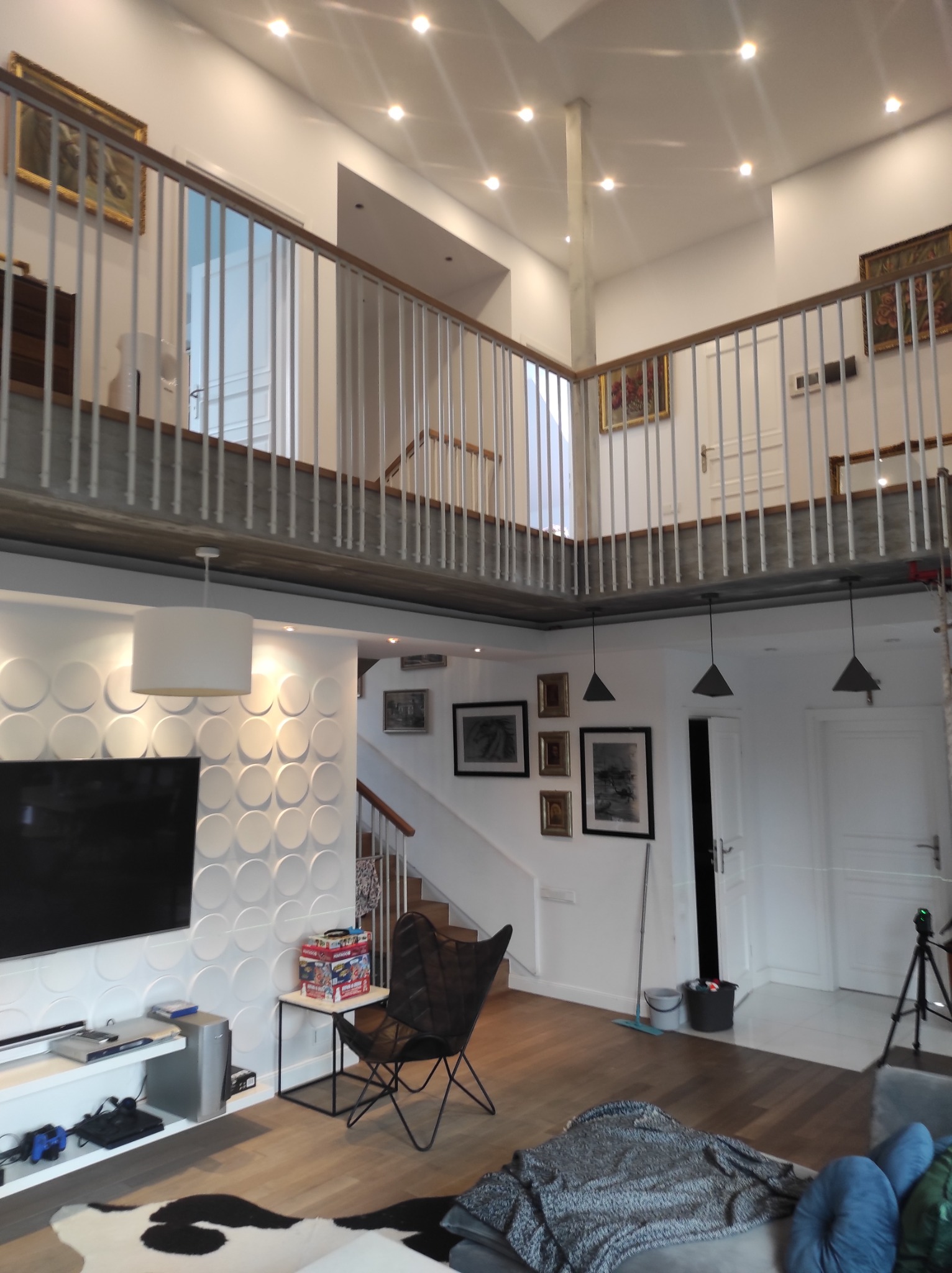 Dwupoziomowy salon z antresolą, białe ściany z dekoracyjnymi panelami 3D, drewniana podłoga, czarne lampy wiszące, galeria obrazów na ścianach, nowoczesne meble.