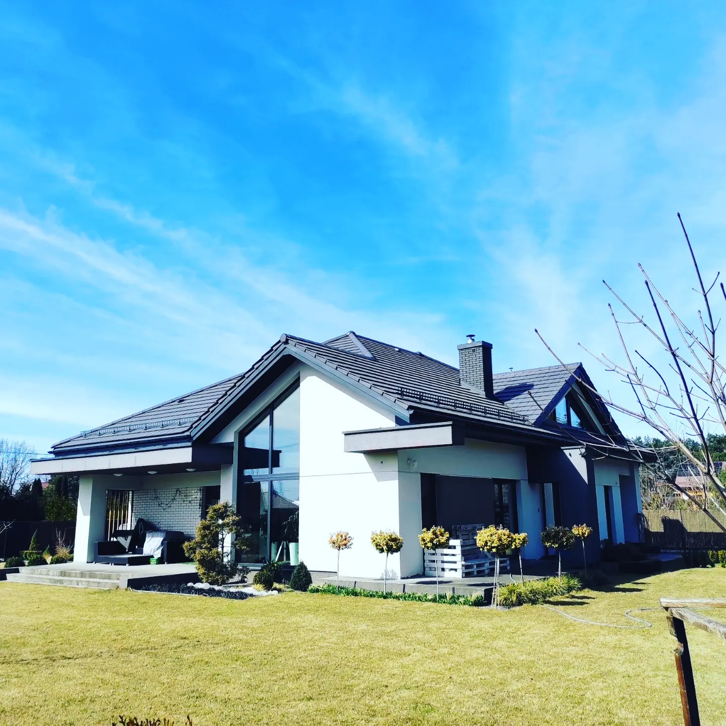 Nowoczesny dom jednorodzinny z dwuspadowym dachem, dużymi przeszkleniami i starannie wypielęgnowanym trawnikiem w słoneczny dzień.