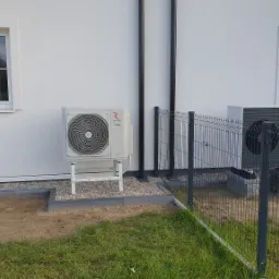 Dwie jednostki zewnętrzne, Rotenso i Vaillant, zainstalowane przy białej ścianie domu, widoczne przez metalowe ogrodzenie, z oknami w tle.
