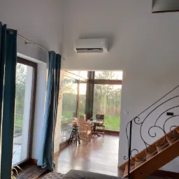 Wnętrze domu z klimatyzatorem ściennym, widok na przeszkloną werandę z wiklinowym fotelem i drewniane schody z ozdobną, kutą balustradą.