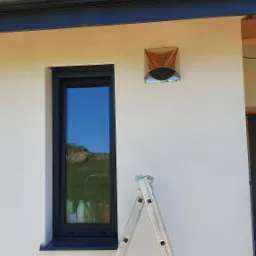 Elewacja domu z zainstalowanym anemostatem wentylacyjnym, widoczna drabina oparta o ścianę, okno z odbiciem krajobrazu.