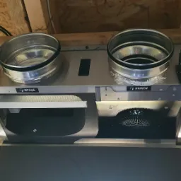 Widok od dołu na zamontowaną centralę wentylacyjną z dwoma króćcami przyłączeniowymi, widocznymi filtrami i wentylatorem, umieszczoną pod drewnianym stropem poddasza, z podłączonym przewodem...