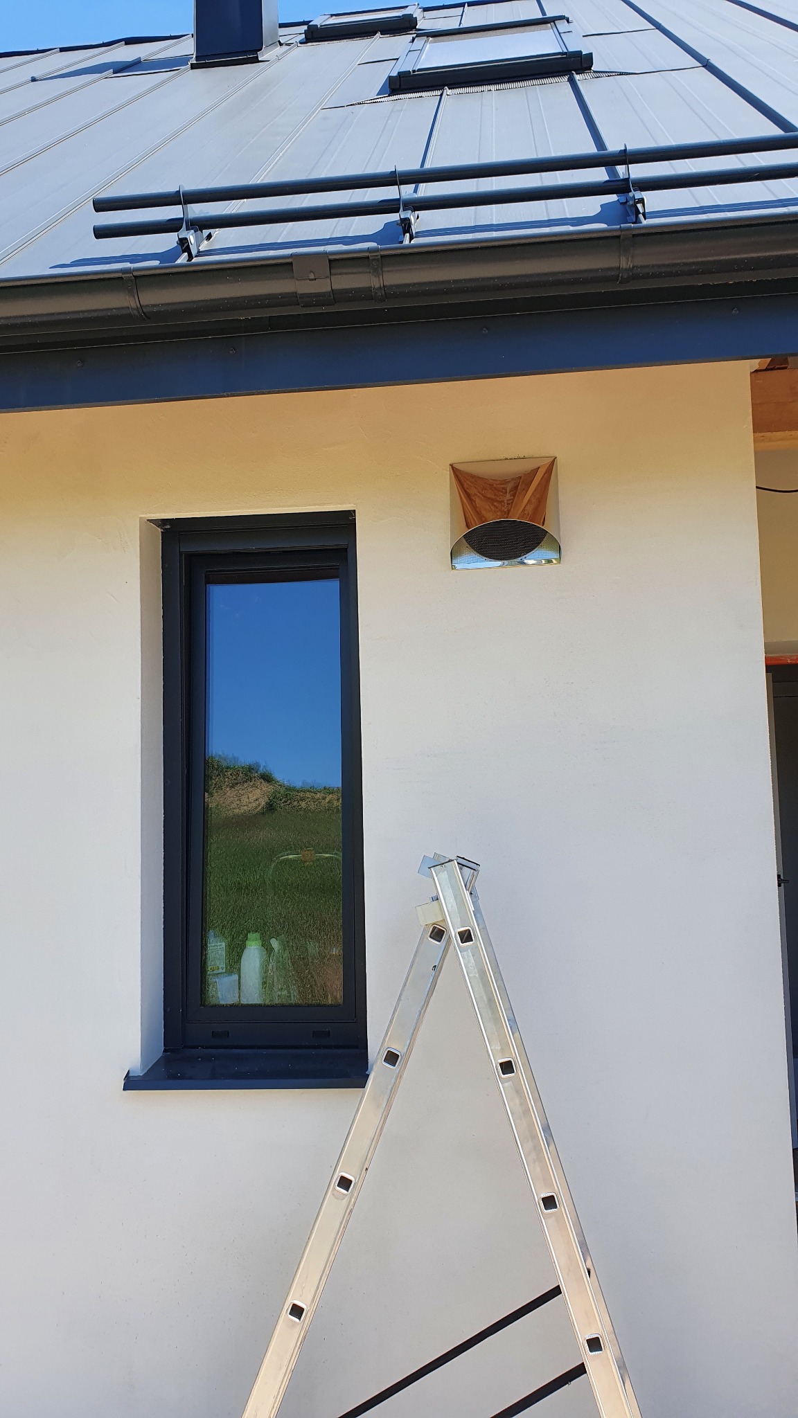 Elewacja domu z zainstalowanym anemostatem wentylacyjnym, widoczna drabina oparta o ścianę, okno z odbiciem krajobrazu.
