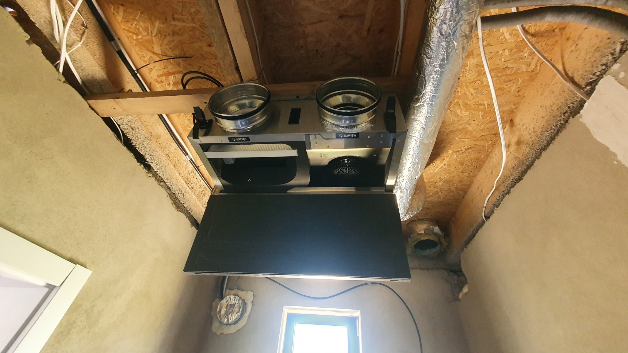 Centrala wentylacyjna z rekuperacją zamontowana pod stropem, widoczne kanały wentylacyjne, otwór okienny w tle.