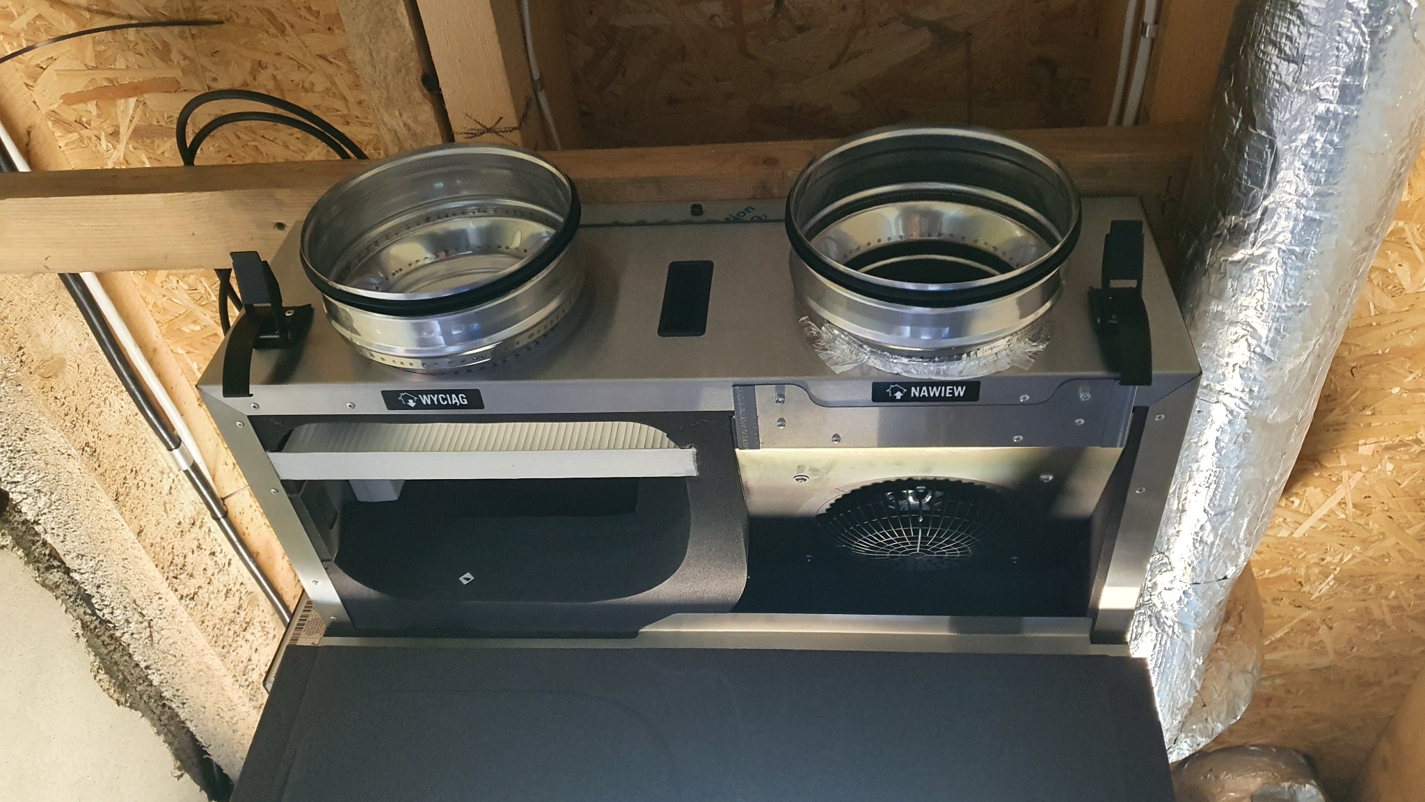 Widok od dołu na zamontowaną centralę wentylacyjną z dwoma króćcami przyłączeniowymi, widocznymi filtrami i wentylatorem, umieszczoną pod drewnianym stropem poddasza, z podłączonym przewodem...