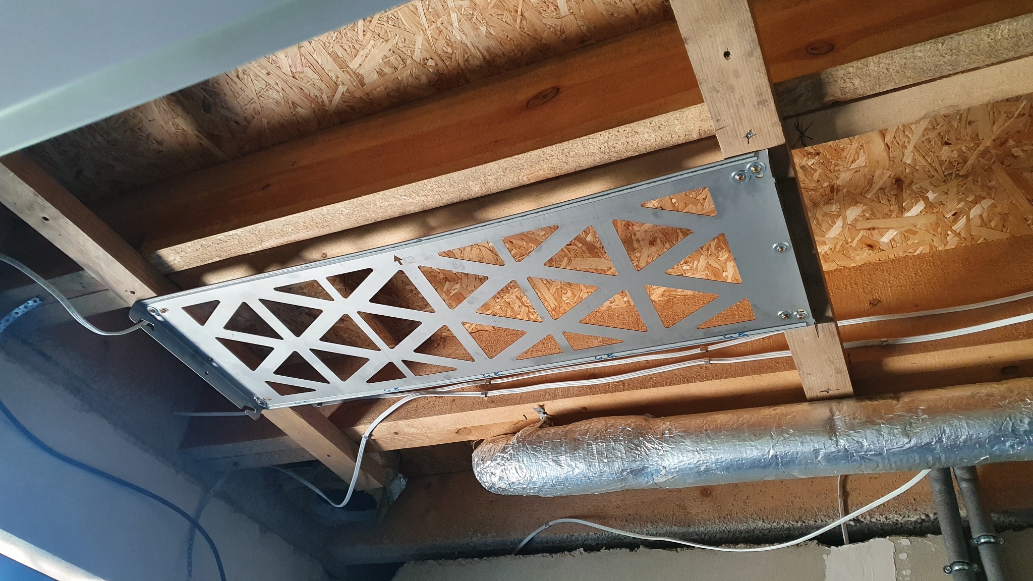 Montaż metalowej kratki wentylacyjnej z trójkątnymi otworami do drewnianej konstrukcji sufitu podwieszanego, widoczne przewody i rura wentylacyjna w folii aluminiowej.