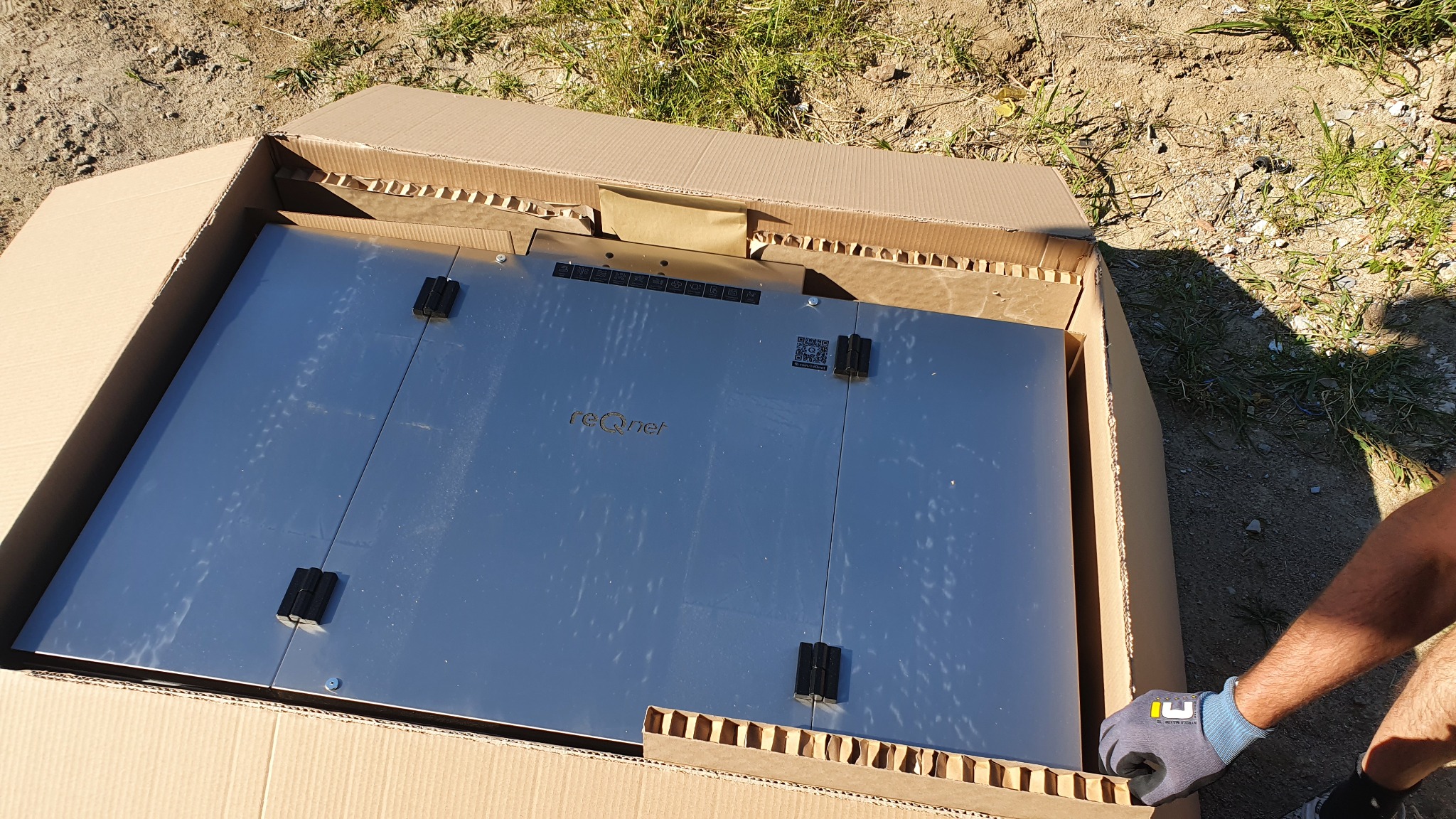 Rozpakowywanie srebrnej centrali wentylacyjnej z widocznym logo 'reQnet' i kodem QR w kartonowym opakowaniu na zewnątrz, widoczna dłoń w rękawicy roboczej.