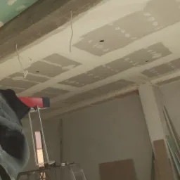 Montaż sufitu podwieszanego z płyt karton-gips, widoczne płyty z wyciętymi otworami na oświetlenie, surowa belka konstrukcyjna, drabina rozstawiona w pomieszczeniu w trakcie prac wykończeniowych.