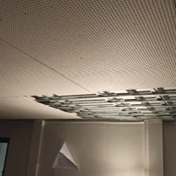 Demontaż perforowanego sufitu podwieszanego z widoczną konstrukcją metalową i częściowo usuniętą płytą, oświetlony lampą roboczą na drabinie, z widocznym planem na ścianie.