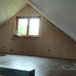 Poddasze w trakcie wykańczania: położone panele podłogowe, boazeria na ścianie pod oknem, płyta karton gips po lewej stronie, widoczne kable i rury od odkurzacza