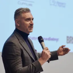 Mężczyzna w garniturze z mikrofonem na tle ekranu z napisem BRIEF podczas prezentacji.