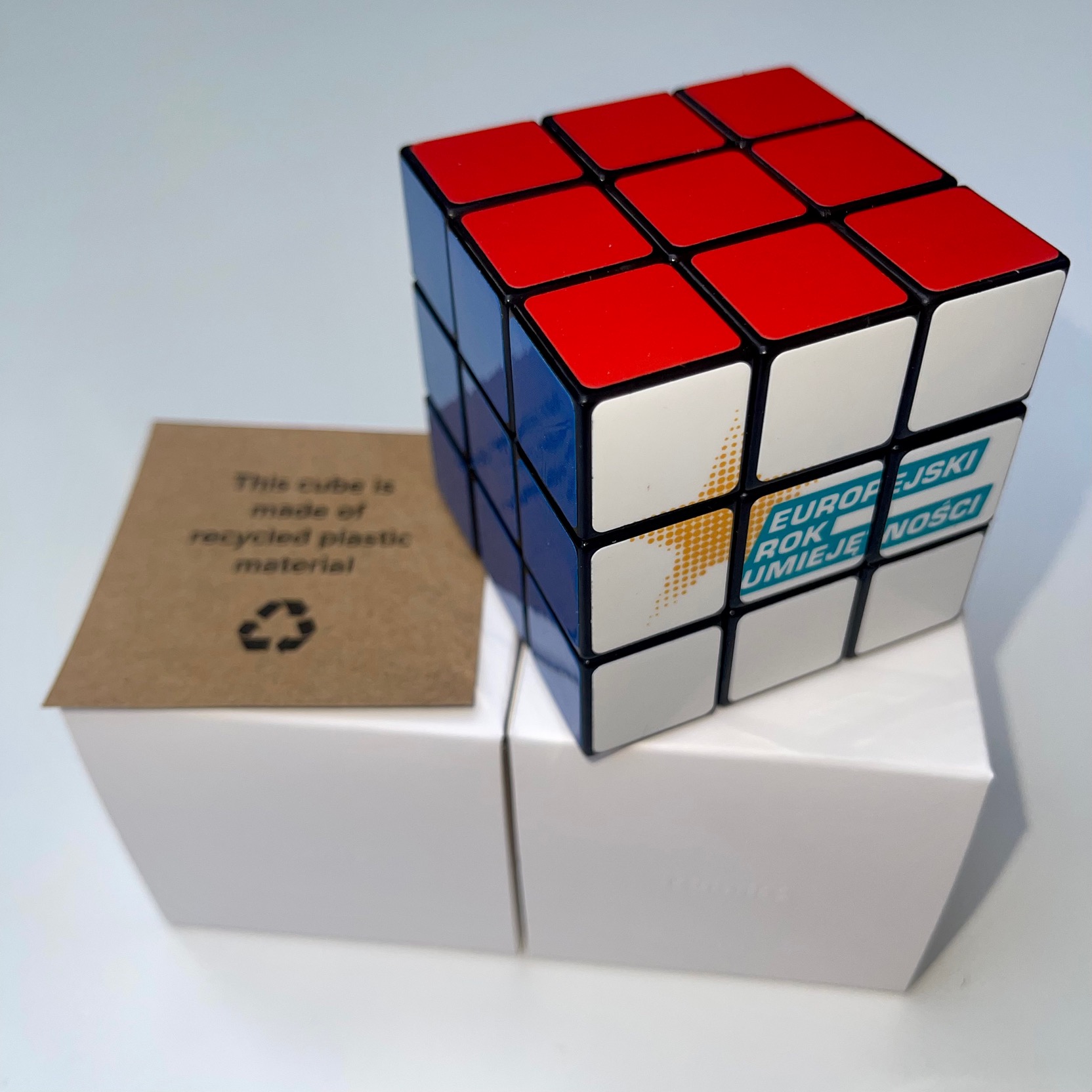 Kostka Rubika z nadrukiem 'Europejski Rok Umiejętności' stojąca na białych pudełkach, obok kartonowa etykieta z informacją o recyklingu.