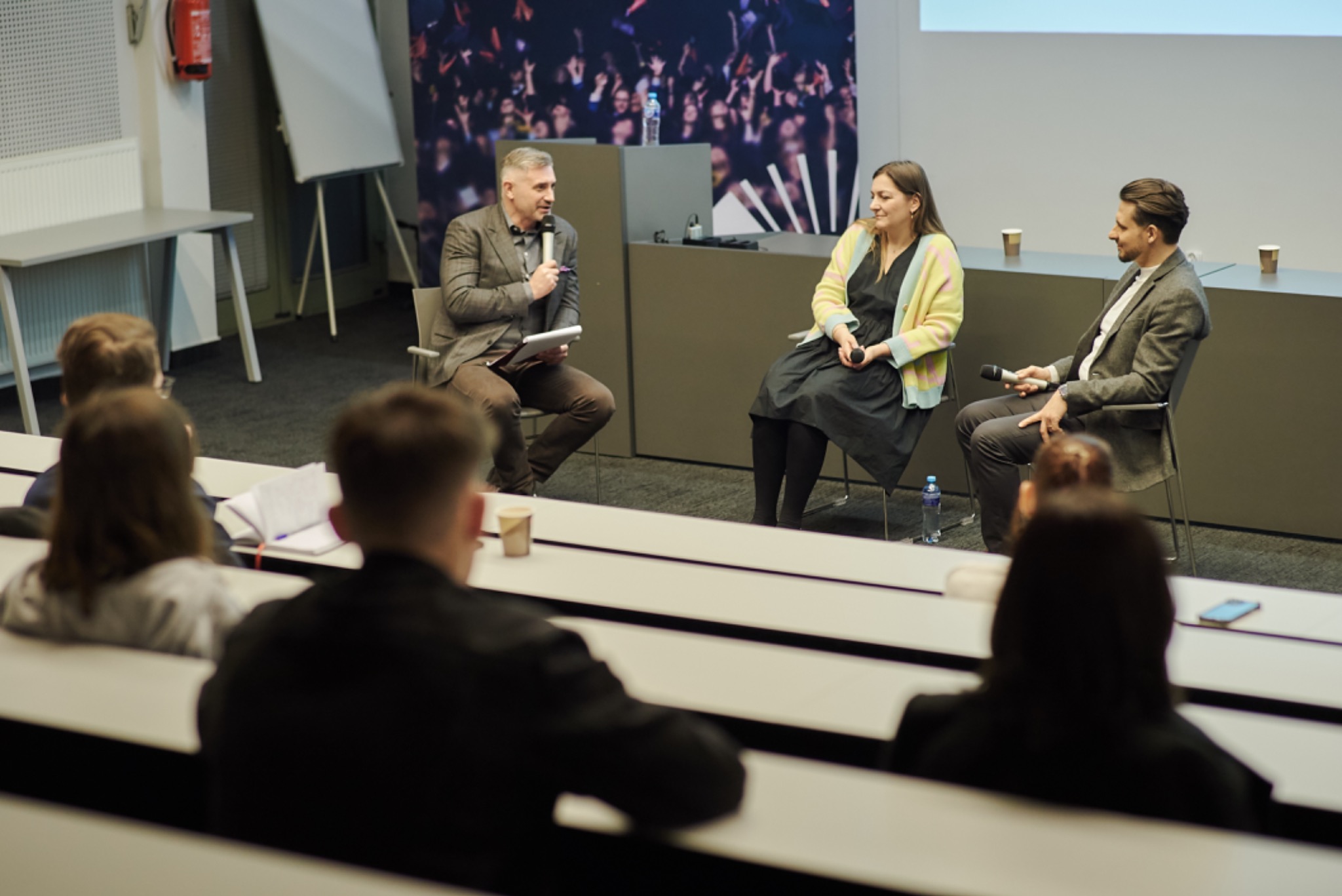 Panel dyskusyjny z trzema prelegentami siedzącymi na krzesłach na scenie przed publicznością w auli. Mężczyzna w brązowej marynarce trzyma mikrofon i tablet, kobieta w kolorowym kardiganie...