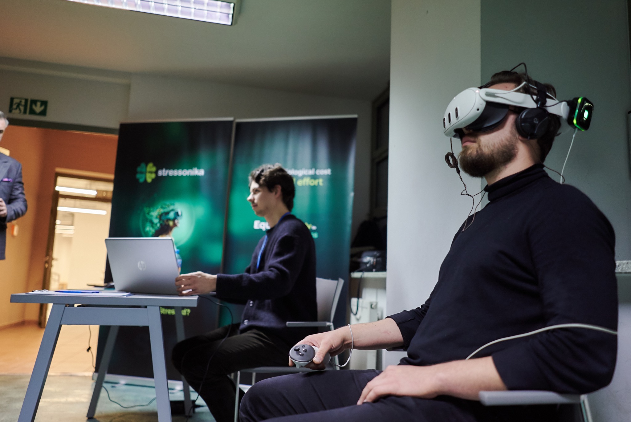 Mężczyzna z brodą używa zestawu VR z kontrolerem; w tle osoba pracuje na laptopie przed banerem reklamowym.