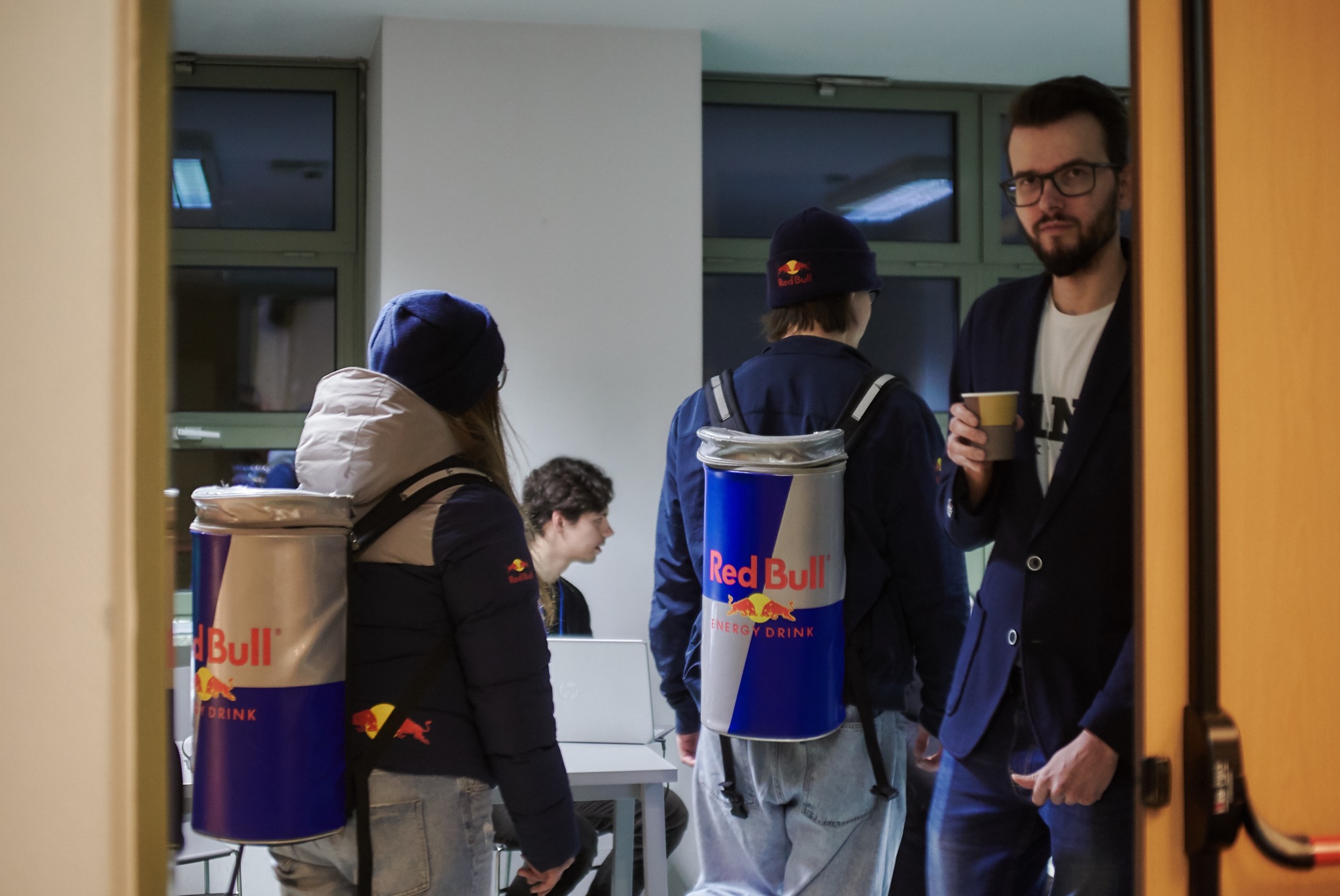 Osoby z plecakami-chłodziarkami Red Bull w pomieszczeniu biurowym; mężczyzna w marynarce trzyma papierowy kubek.
