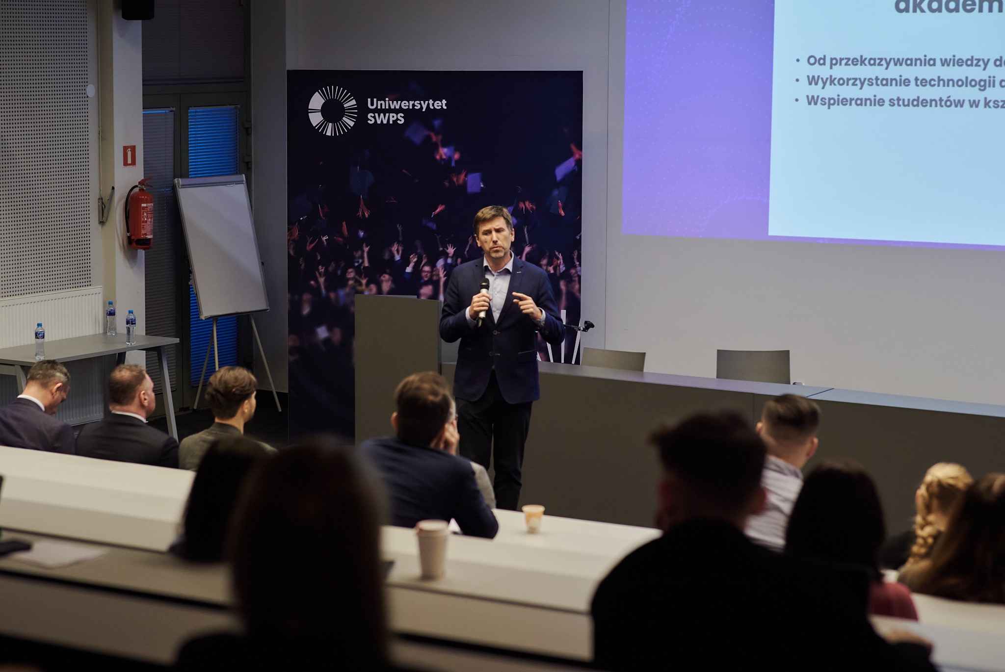 Prelegent w ciemnym garniturze przemawia do publiczności w auli uniwersyteckiej, z logo Uniwersytetu SWPS widocznym na tle.