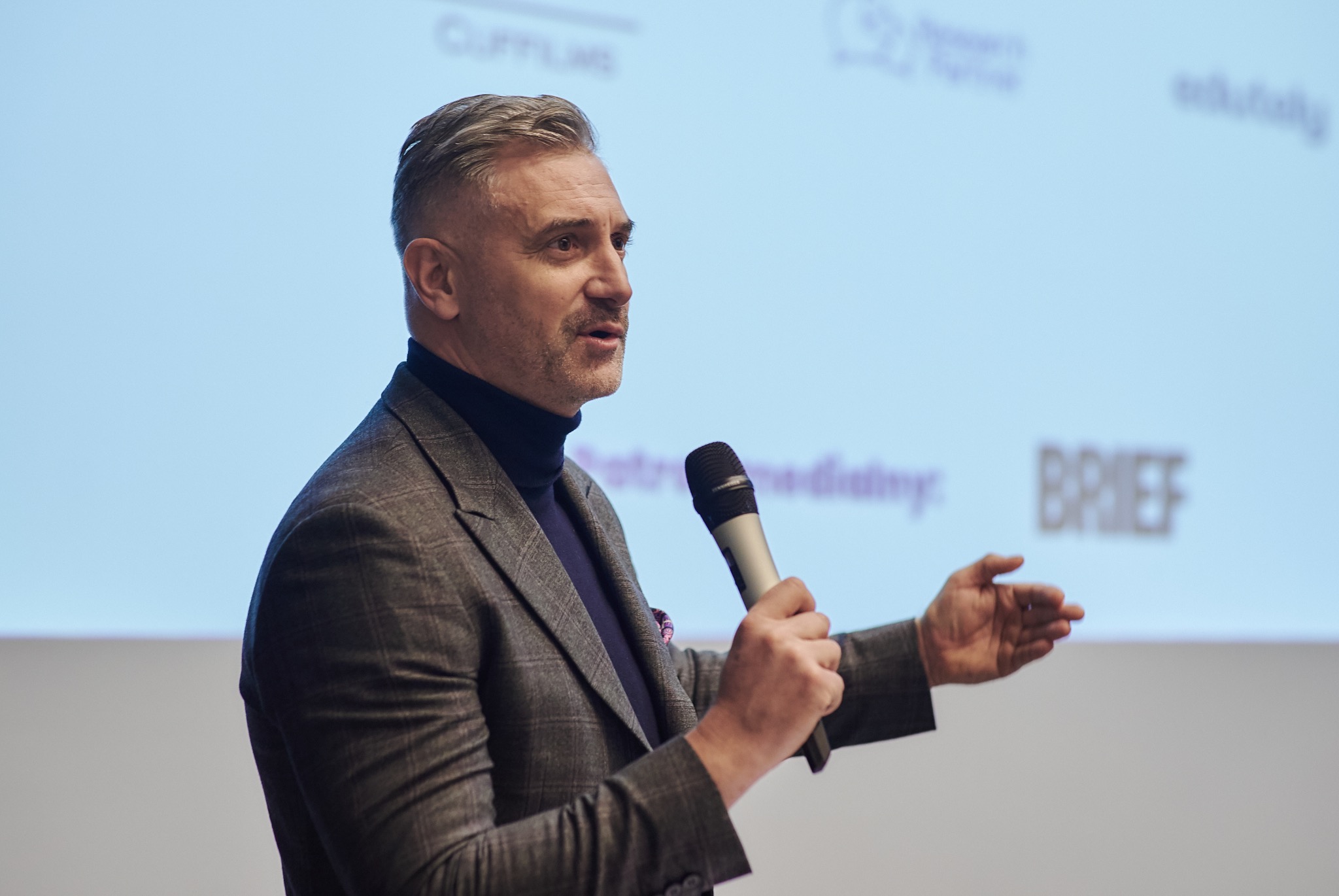 Mężczyzna w garniturze z mikrofonem na tle ekranu z napisem BRIEF podczas prezentacji.