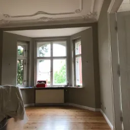 Pokój po malowaniu w Tczewie, z widoczną drewnianą podłogą, szarymi ścianami, ozdobnym sufitem i oknem wykuszowym z widokiem na zieleń.