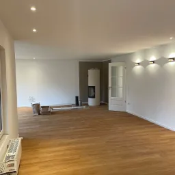 Jasne panele podłogowe w przestronnym, odnowionym salonie z białymi ścianami, nowoczesnym kominkiem i trzema ściennymi lampami z ciepłym światłem. Częściowo rozpakowane materiały budowlane w rogu...