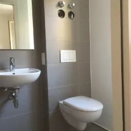 Ukończona łazienka w szarych płytkach z wiszącą toaletą, umywalką i niedokończonymi wylewkami baterii.