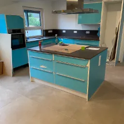 Montaż kuchni: turkusowe szafki z brązowym blatem, wyspa kuchenna z narzędziami i folią ochronną, okap AEG, etap wykończeniowy.