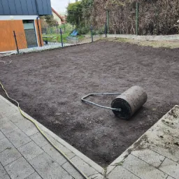 Przygotowany teren pod trawnik z ubitą glebą i walcem ogrodowym, obok zwinięty wąż ogrodowy, widok z perspektywy chodnika z kostki brukowej.