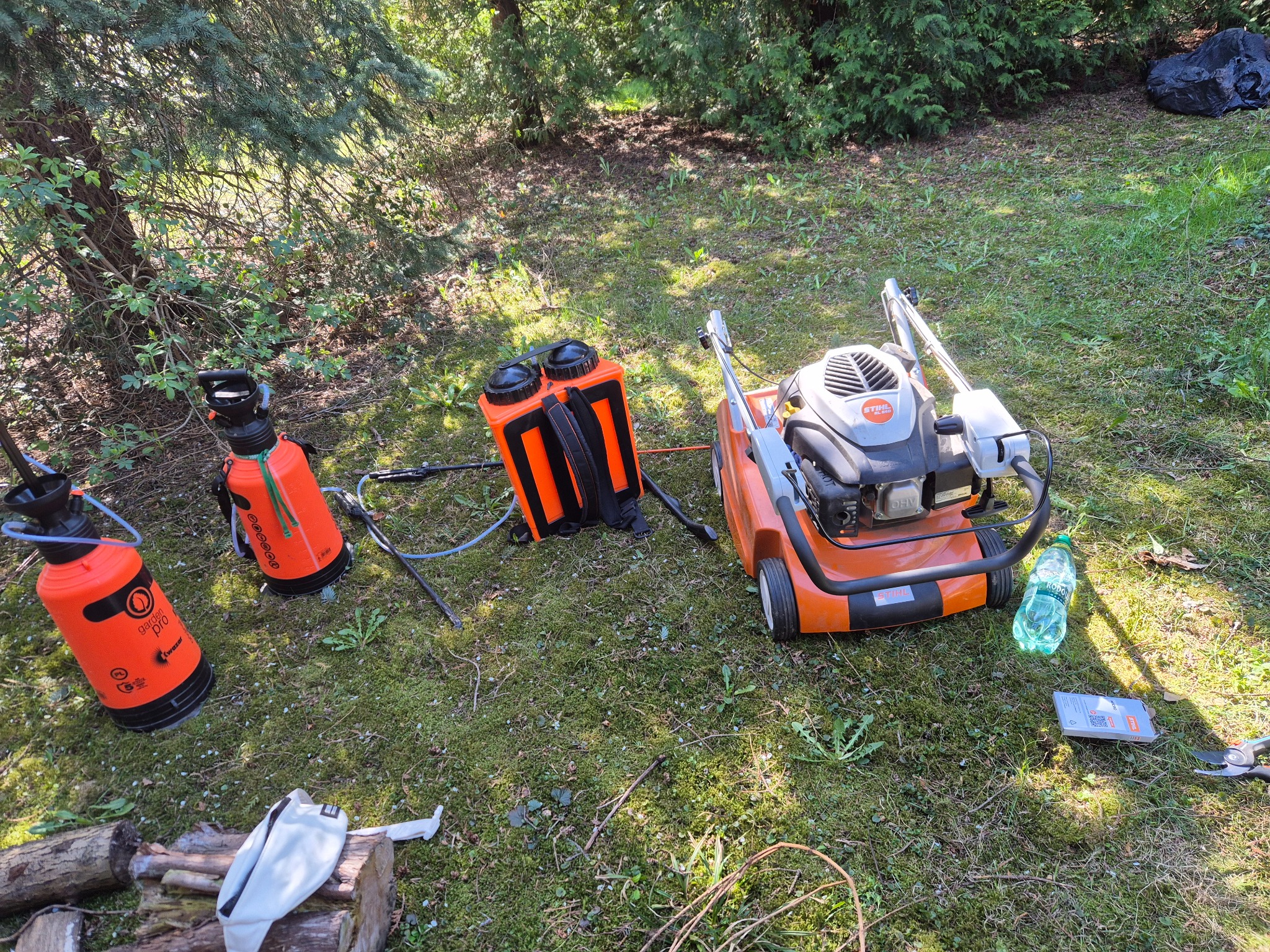 Sprzęt ogrodniczy: kosiarka Stihl, opryskiwacze ciśnieniowe Garden Pro Kwazar, opryskiwacz plecakowy, sekator, woda i akcesoria na trawie w ogrodzie.