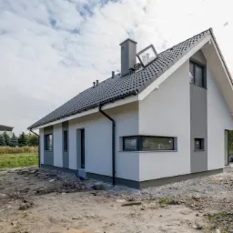 Nowoczesny dom jednorodzinny z dwuspadowym dachem pokrytym grafitową dachówką, z oknem dachowym uchylonym do góry, elewacja w odcieniach bieli i szarości, widoczny fragment terenu przed budynkiem...