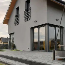 Szary dom jednorodzinny z płaskim dachem i dużymi oknami narożnymi, widoczne metalowe balustrady przy oknach na piętrze, otoczenie domu wyłożone szarą kostką brukową, widoczne fragmenty trawnika.