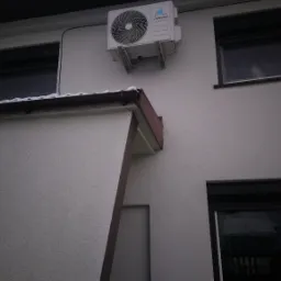 Biała jednostka zewnętrzna klimatyzacji Vesser zamontowana na elewacji budynku z oknami w zimowej scenerii, widoczne opady śniegu.