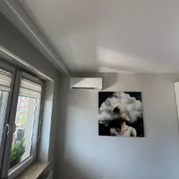 Biały klimatyzator ścienny zamontowany nad obrazem w nowoczesnym mieszkaniu z oknem balkonowym.